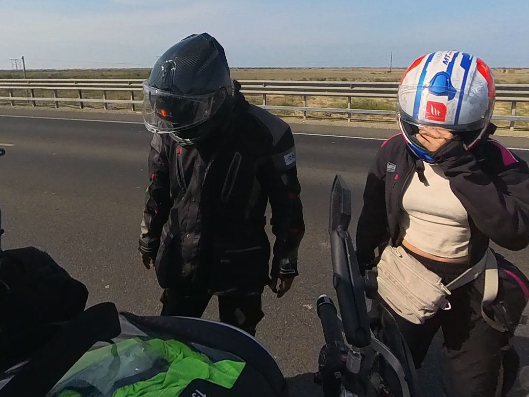 Dos viajeros con casco junto a la moto al borde de la carretera.